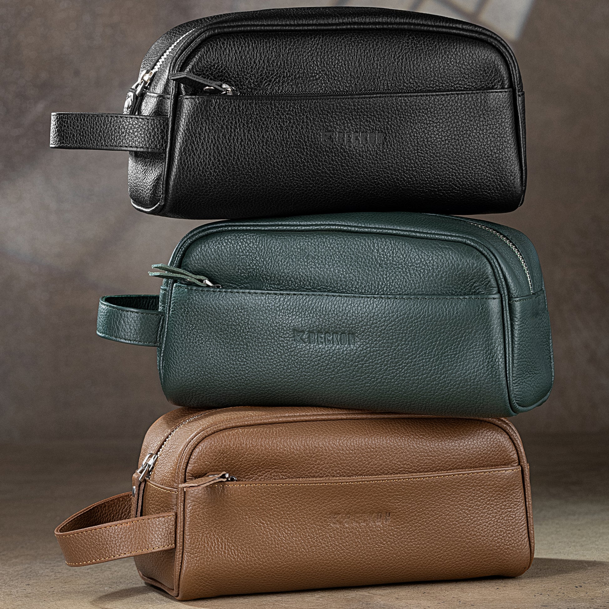 Three leather toiletry bags in black, green, and brown on top of each other
